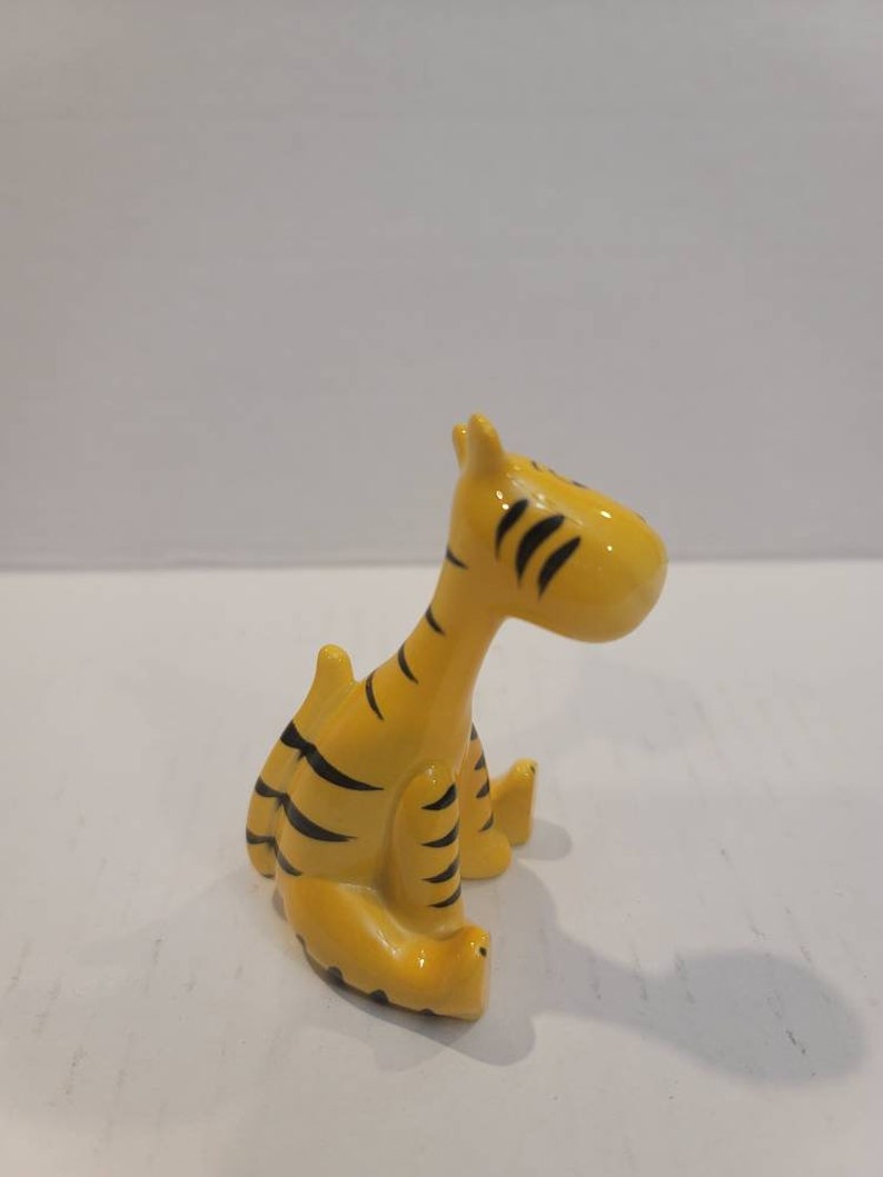 May include: A small ceramic figurine of a tiger sitting down. The tiger is yellow with black stripes and has a simple, cartoon-like design.