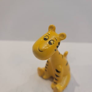May include: A small yellow ceramic figurine of a tiger with black stripes. The tiger is sitting with its head tilted to the side and has a smiling expression.