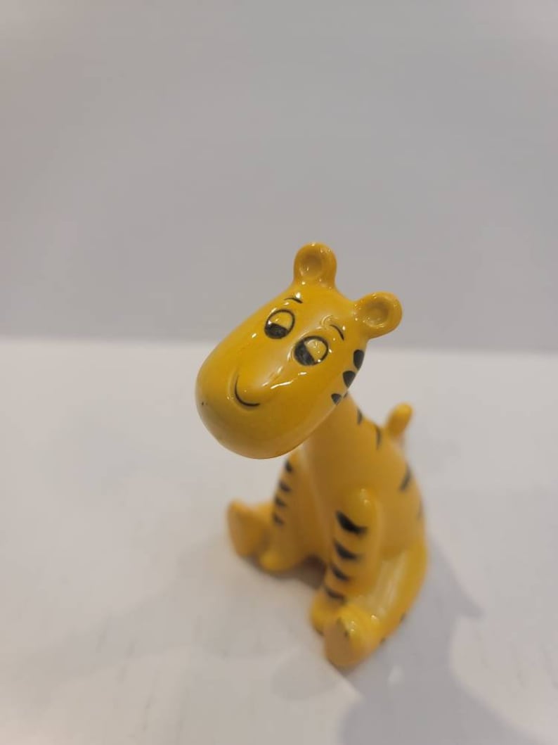 May include: A small yellow ceramic figurine of a tiger with black stripes. The tiger is sitting with its head tilted to the side and has a smiling expression.