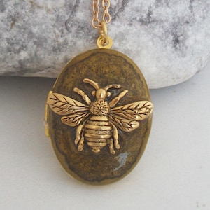 May include: Gold-coloured bee pendant necklace. The oval pendant features a detailed bee design on a textured, olive-green background. The necklace has a delicate gold chain, suitable for everyday wear.