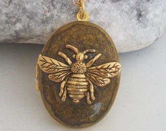May include: Gold-toned bee pendant necklace. The oval pendant features a detailed bee design on a textured, olive-green background. The necklace has a delicate gold chain, perfect for everyday wear.
