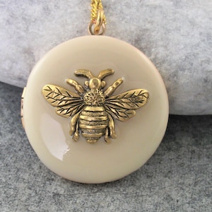 May include: A cream-coloured round pendant necklace featuring a gold-coloured bee charm. The bee has detailed, textured wings and body. The pendant hangs from a delicate gold chain, creating a classic and elegant accessory.