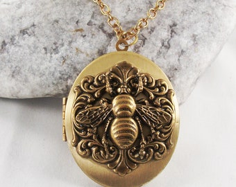 May include: An oval-shaped brass-toned metal locket pendant featuring a raised relief design of a bee centered within ornate scrollwork. The locket is attached to a matching gold-toned link chain, showcasing a textured finish and a hinged side closure.