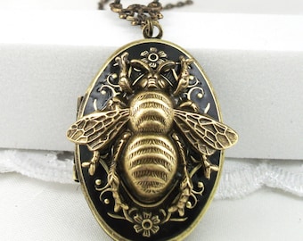 May include: A gold bee locket necklace with a black enamel background. The bee is detailed with raised wings and a textured body. The locket is oval shaped and has a delicate chain.