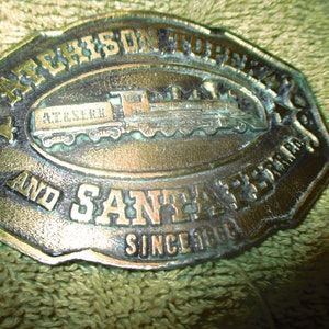 May include: A brass belt buckle with a raised design of a train engine and the text "Atchison, Topeka and Santa Fe Railroad Since 1880".