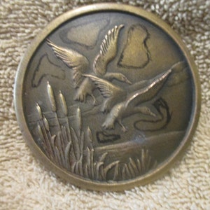 Pode incluir: Uma fivela de cinto de bronze com um design detalhado de três patos voando sobre um pântano com juncos. Os patos estão em voo e os juncos estão em primeiro plano.