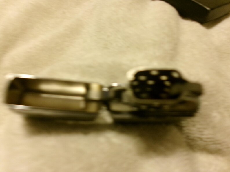 May include: A silver-toned metal lighter, open to reveal the interior. The lighter's top section is black with white polka dots. The lighter is on a light-colored, textured surface.