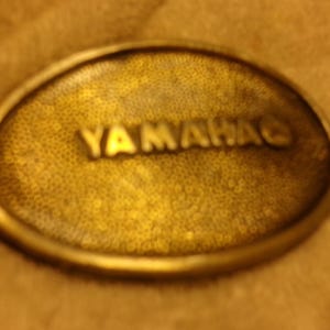 May include: Oval-shaped, antique brass-colored belt buckle with the word "YAMAHA" embossed in raised lettering. The buckle has a textured surface and a rounded edge, suggesting a vintage or retro style.