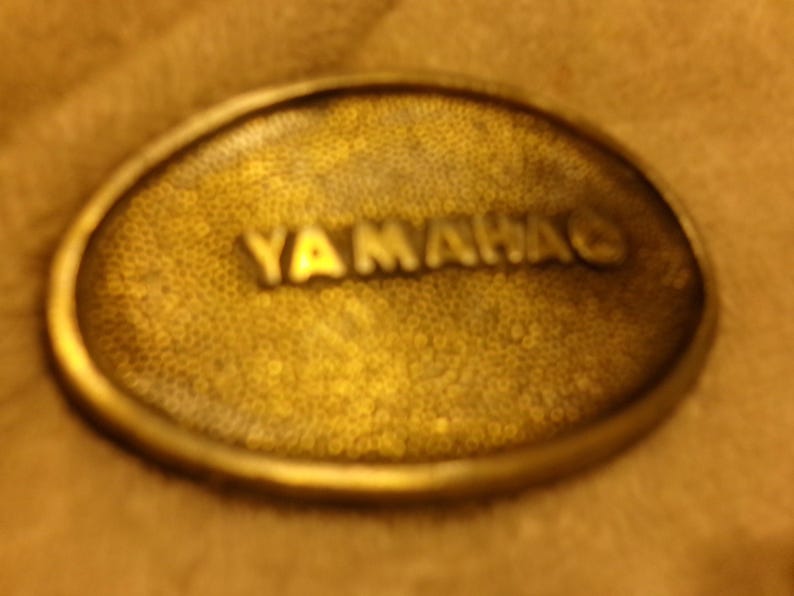 May include: Oval-shaped, antique brass-colored belt buckle with the word "YAMAHA" embossed in raised lettering. The buckle has a textured surface and a rounded edge, suggesting a vintage or retro style.