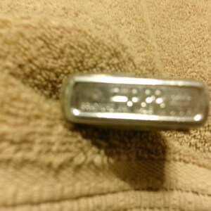 May include: A silver-colored lighter with text on the front. The lighter is rectangular with rounded edges. The lighter is resting on a tan, textured surface.