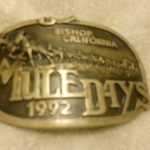 May include: A gold-toned metal belt buckle with the text "Bishop California Mule Days 1992" and a depiction of a mule pulling a cart.