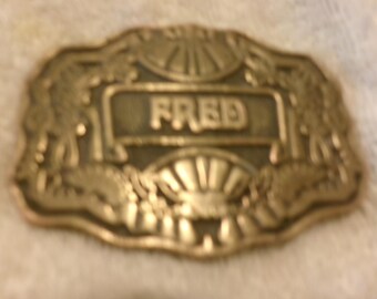 Könnte beinhalten: Eine goldfarbene Gürtelschnalle im Vintage-Stil mit dem Namen "FRED" in der Mitte. Die Schnalle hat einen verzierten Rand mit Sonnenstrahlen- und blütenartigen Designs. Die Form ist rechteckig mit abgerundeten Kanten.