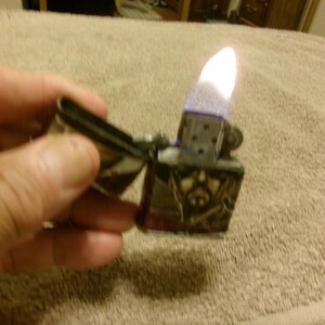 May include: A silver metal lighter with a skull design is open and lit, showing a bright flame. The lighter is held in a hand against a neutral-colored background.
