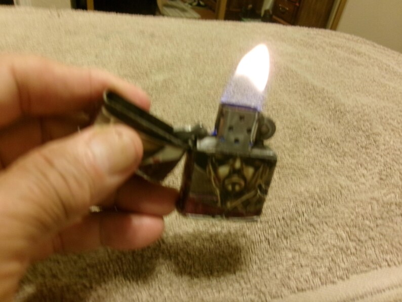 May include: A silver metal lighter with a skull design is open and lit, showing a bright flame. The lighter is held in a hand against a neutral-colored background.