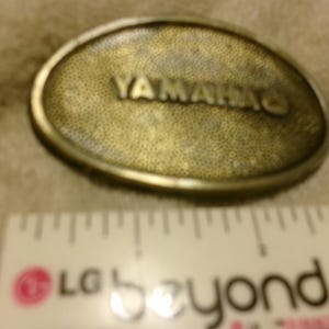 May include: Oval-shaped, brass-colored belt buckle with the word "YAMAHA" in raised lettering. The buckle has a textured surface and a metallic border. A ruler is in the foreground.