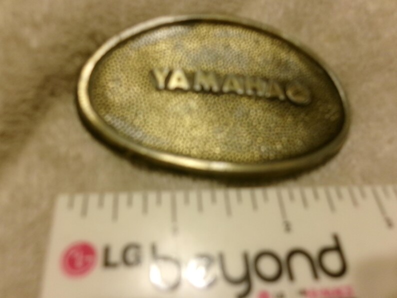 May include: Oval-shaped, brass-colored belt buckle with the word "YAMAHA" in raised lettering. The buckle has a textured surface and a metallic border. A ruler is in the foreground.
