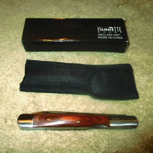 May include: A brown wooden handled pocket knife with a silver blade. The knife is in a black pouch and comes in a black box with the text "HAMMETTE  SHARP (883) 299-3901 MADE IN CHINA".