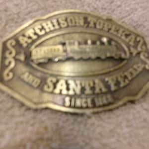 May include: A gold-colored metal belt buckle with the text "Atchison, Topeka and Santa Fe Since 1869" and a depiction of a train.