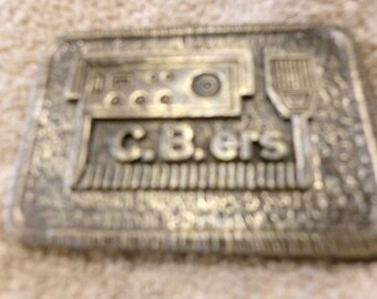 Könnte beinhalten: Silberfarbener Gürtelschnalle aus Metall mit einem CB-Radio-Design und dem Text "C.B.ers".