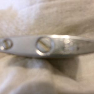 Puede incluir: Un objeto met&aacute;lico plateado y alargado con dos tornillos y una abertura circular. El objeto parece ser un encendedor, con un dise&ntilde;o ligeramente curvado y una superficie lisa y reflectante. El encendedor est&aacute; sobre una tela blanca texturizada.