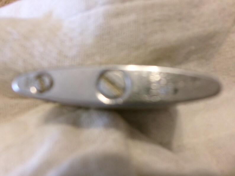 Puede incluir: Un objeto met&aacute;lico plateado y alargado con dos tornillos y una abertura circular. El objeto parece ser un encendedor, con un dise&ntilde;o ligeramente curvado y una superficie lisa y reflectante. El encendedor est&aacute; sobre una tela blanca texturizada.