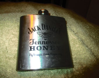 May include: A silver metal flask with a black Jack Daniel's Tennessee Honey logo and a bee design. The text "Fly Straight. Drink Responsibly" is printed below the logo.