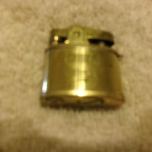 May include: A gold-colored metal lighter with a simple design. The lighter has a rounded shape and a smooth surface.