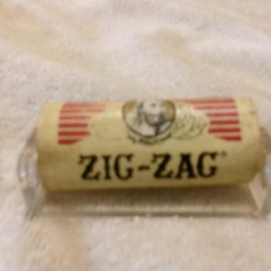 May include: A Zig-Zag rolling paper dispenser with a cream-colored paper roll. The roll has red stripes and the Zig-Zag logo. The dispenser is clear plastic.