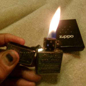 May include: A lit Zippo lighter with a flame, held open. The lighter is silver with engraved details. A black Zippo box with the brand name is in the background. The flame is yellow and blue.