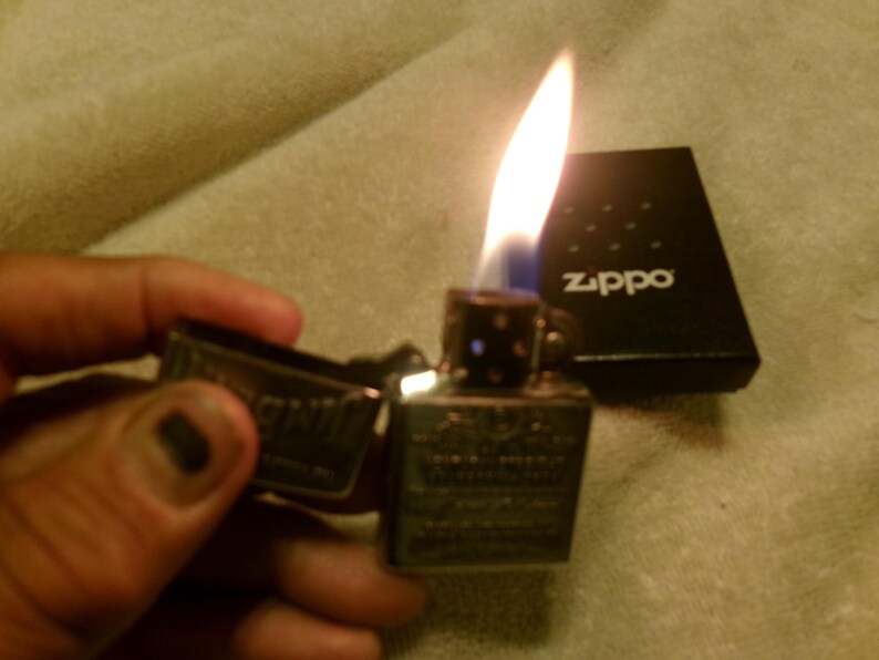 May include: A lit Zippo lighter with a flame, held open. The lighter is silver with engraved details. A black Zippo box with the brand name is in the background. The flame is yellow and blue.