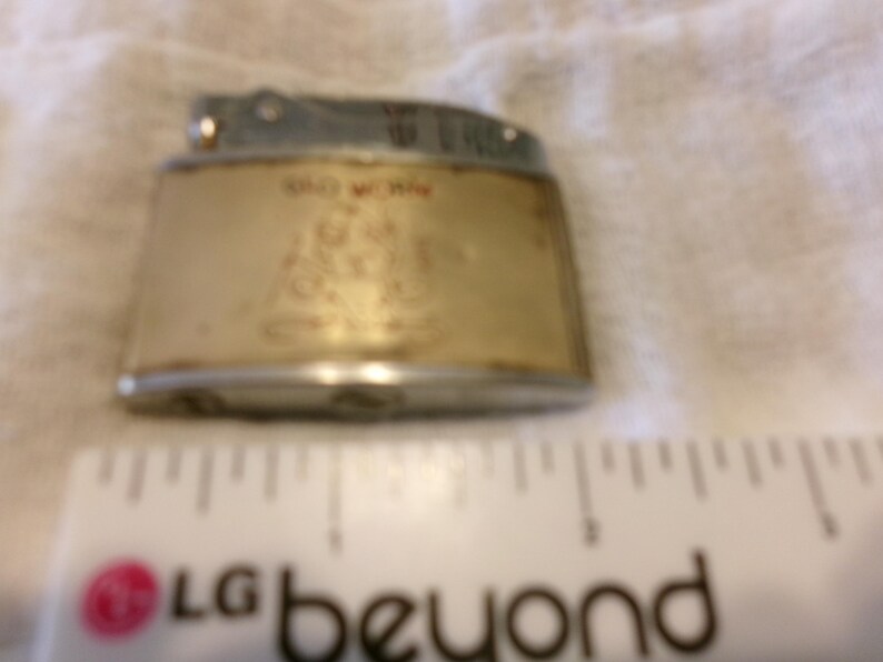 Puede incluir: Encendedor vintage con tapa plateada y cuerpo dorado. El cuerpo tiene las palabras "OLD WORK" y una imagen. El encendedor est&aacute; sobre una superficie blanca.