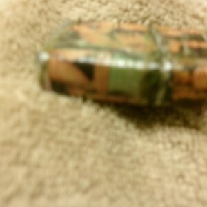 May include: A camouflage-patterned lighter with shades of green, brown, and orange. The lighter is rectangular and appears to be made of metal or a similar material. The image is taken from a slightly elevated angle, showing the top and side of the lighter.