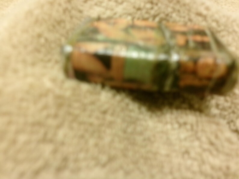 May include: A camouflage-patterned lighter with shades of green, brown, and orange. The lighter is rectangular and appears to be made of metal or a similar material. The image is taken from a slightly elevated angle, showing the top and side of the lighter.