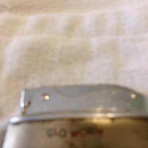 Puede incluir: Un encendedor vintage con la parte superior plateada y una base dorada. El texto "Glo Worn" es visible en letras rojas en la base. El encendedor parece estar usado, mostrando signos de desgaste.