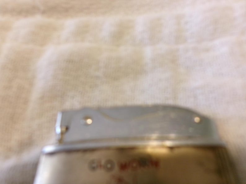 Puede incluir: Un encendedor vintage con la parte superior plateada y una base dorada. El texto "Glo Worn" es visible en letras rojas en la base. El encendedor parece estar usado, mostrando signos de desgaste.