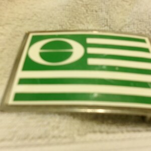 Könnte beinhalten: Rechteckige Metallschnalle mit grün-weißem Design. Das Design zeigt einen weißen Buchstaben "O" mit einem horizontalen Strich und weiße horizontale Streifen auf grünem Hintergrund.