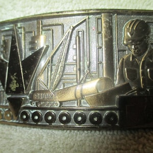 Puede incluir: Una hebilla de cinturón de metal dorado con una escultura en relieve detallada de un trabajador de la construcción operando una grúa. La hebilla tiene la palabra "Fluor" grabada en la grúa.