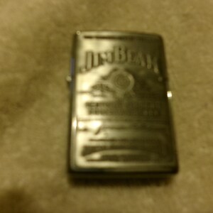 May include: A silver-colored lighter with the "Jim Beam" logo and other text etched on the front. The lighter has a rectangular shape and a hinged lid. The background is a light brown, textured surface.