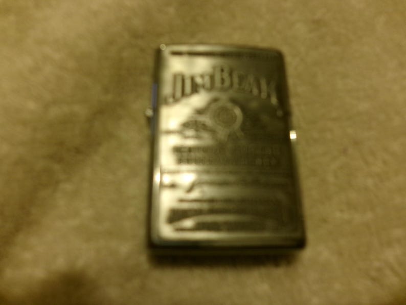 May include: A silver-colored lighter with the "Jim Beam" logo and other text etched on the front. The lighter has a rectangular shape and a hinged lid. The background is a light brown, textured surface.