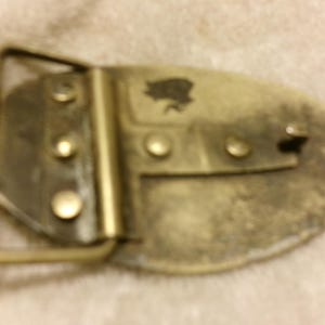 May include: A vintage brass belt buckle with an oval shape. The buckle has a rectangular frame and a decorative emblem. The metal has a worn, aged appearance, with visible rivets and a tarnished finish.