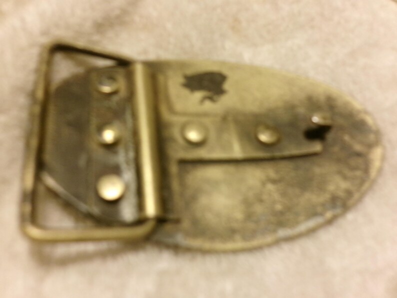 May include: A vintage brass belt buckle with an oval shape. The buckle has a rectangular frame and a decorative emblem. The metal has a worn, aged appearance, with visible rivets and a tarnished finish.