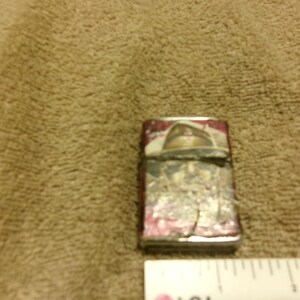 May include: A rectangular, silver-colored lighter with a red and white design on the front. The lighter shows signs of wear and tear, with some of the paint chipped off. The design appears to be a vintage or antique style.