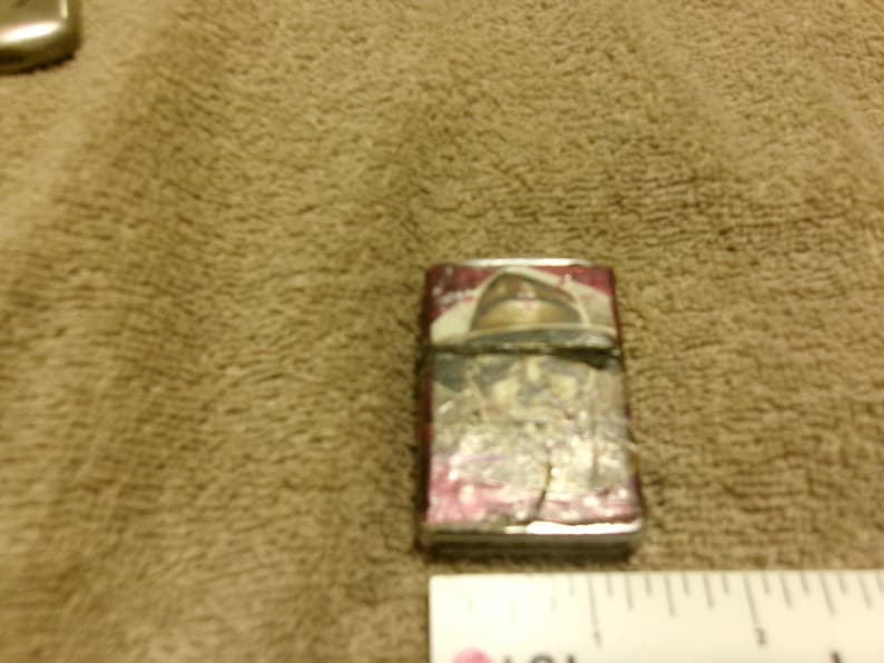 May include: A rectangular, silver-colored lighter with a red and white design on the front. The lighter shows signs of wear and tear, with some of the paint chipped off. The design appears to be a vintage or antique style.