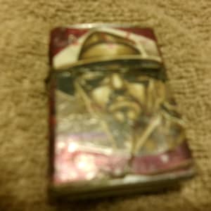 May include: A rectangular lighter with a portrait of a man wearing a hat and sunglasses. The lighter has a red and white background with a silver metal casing. The image is slightly blurry.
