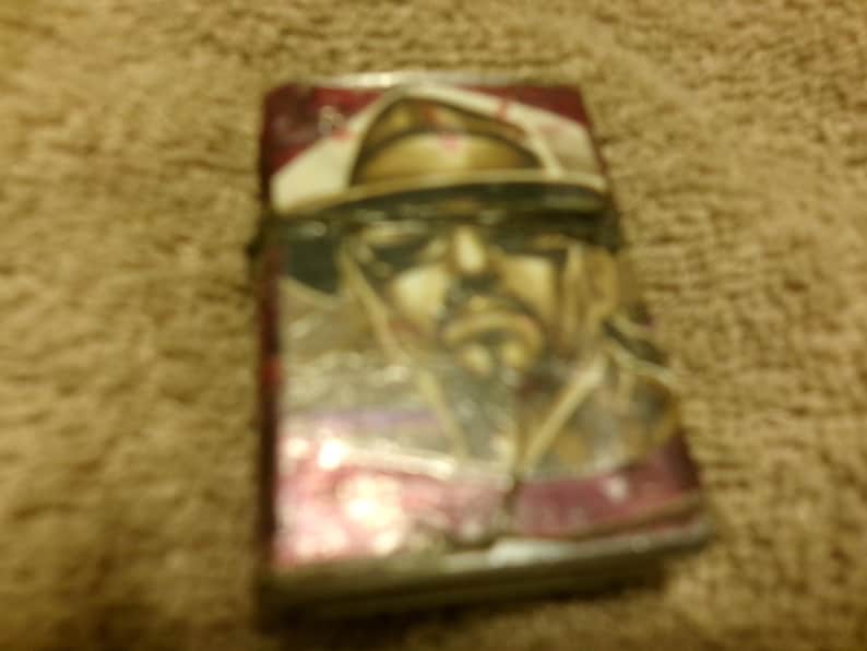 May include: A rectangular lighter with a portrait of a man wearing a hat and sunglasses. The lighter has a red and white background with a silver metal casing. The image is slightly blurry.