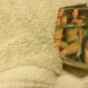 May include: A silver-colored lighter with a printed image of two women in swimwear. The lighter has a rectangular shape and appears to be made of metal. The background is a light beige color.