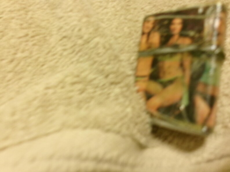 May include: A silver-colored lighter with a printed image of two women in swimwear. The lighter has a rectangular shape and appears to be made of metal. The background is a light beige color.