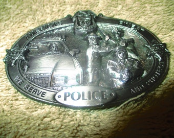 Puede incluir: Una hebilla de cinturón de metal plateado con temática policial. Presenta un coche de policía, tres oficiales y las palabras "American Police We Serve And Protect".