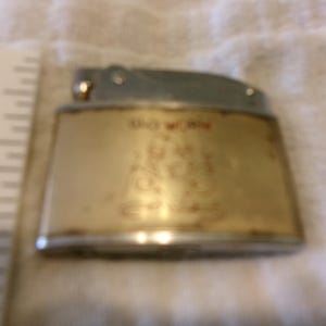 Puede incluir: Un encendedor vintage con una parte superior plateada y un cuerpo dorado. El cuerpo tiene texto rojo que dice "DO WORK" y una ilustraci&oacute;n. El encendedor est&aacute; sobre una superficie blanca.