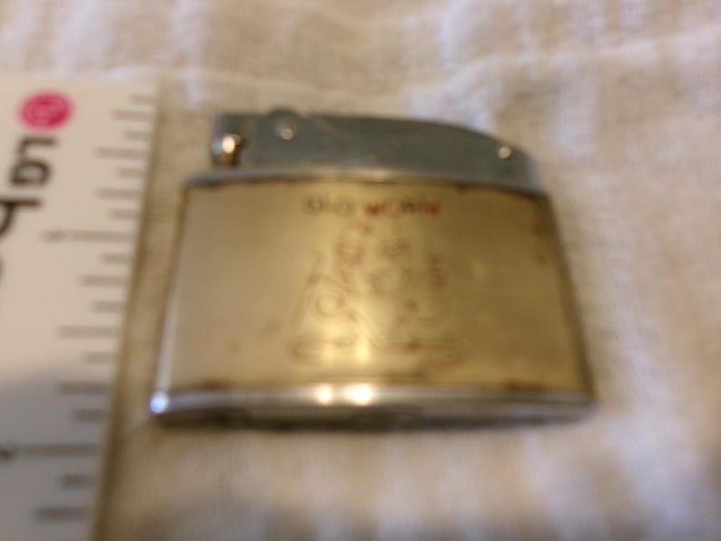 Puede incluir: Un encendedor vintage con una parte superior plateada y un cuerpo dorado. El cuerpo tiene texto rojo que dice "DO WORK" y una ilustraci&oacute;n. El encendedor est&aacute; sobre una superficie blanca.